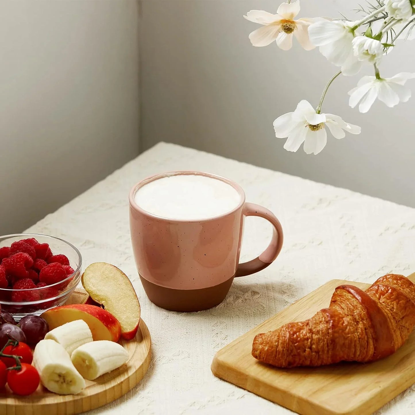 Pink and brown ceramic coffee mug with foam, croissant on wooden board, fresh fruit and flowers