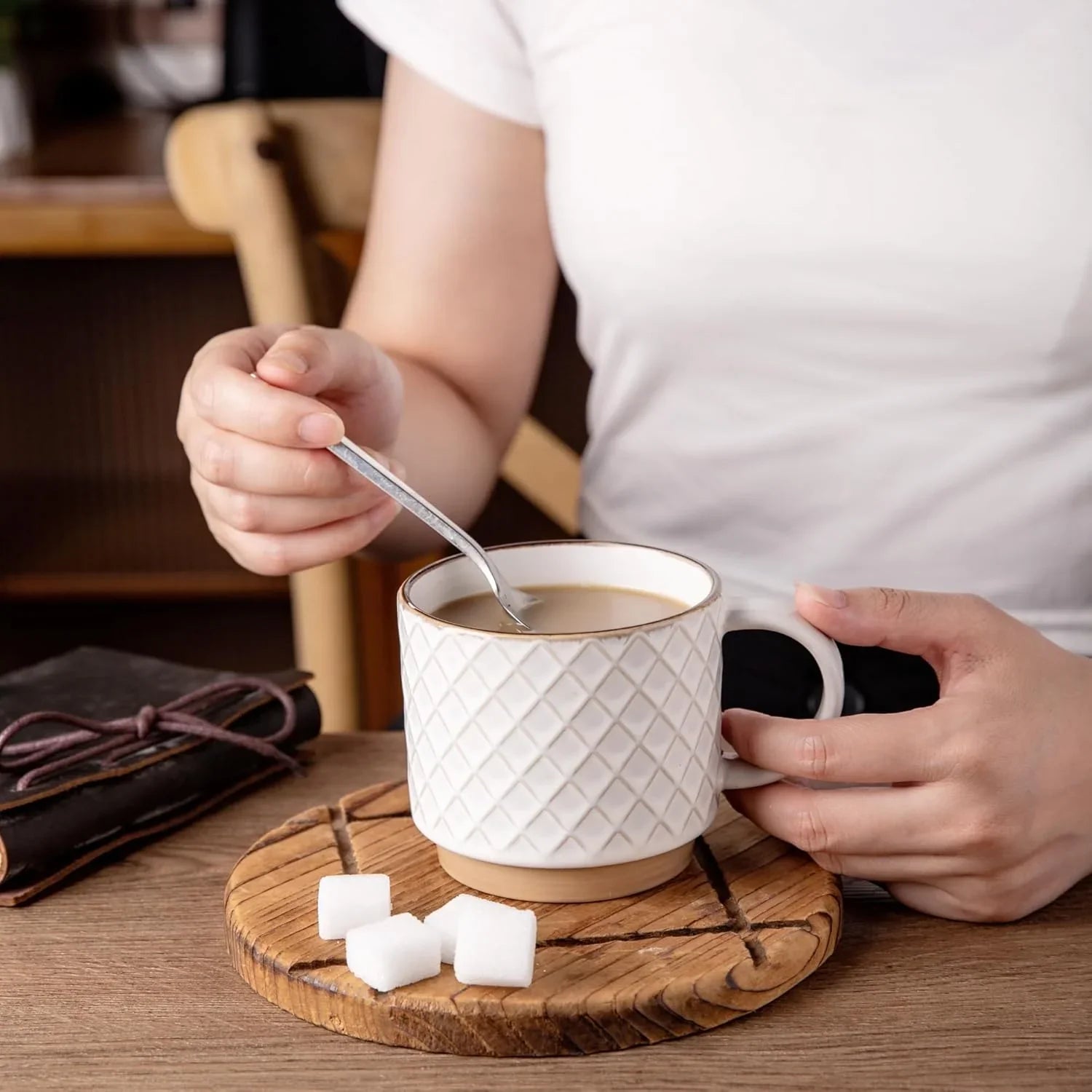 Person stirring coffee in a textured off-white ceramic mug on a wooden coaster with sugar cubes