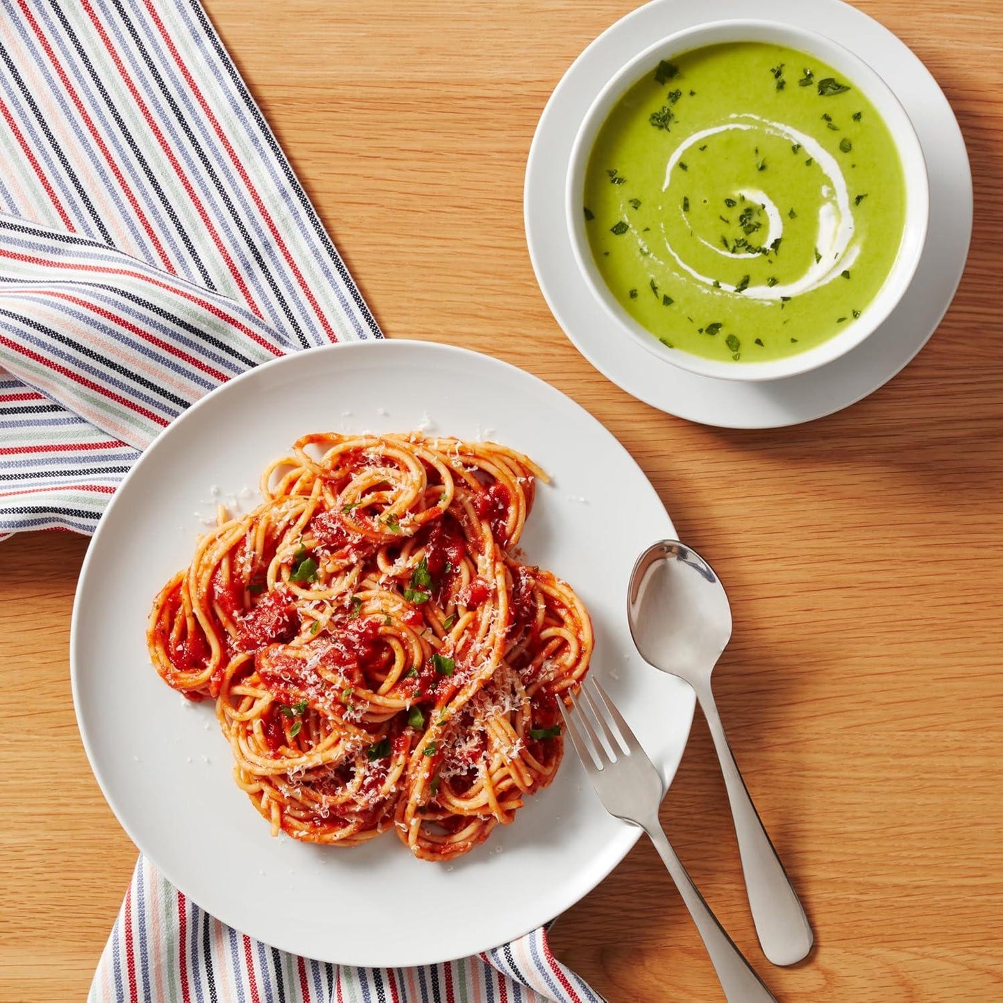 White porcelain plate with spaghetti marinara and grated cheese, bowl of green soup with cream swirl, fork and spoon on wooden table