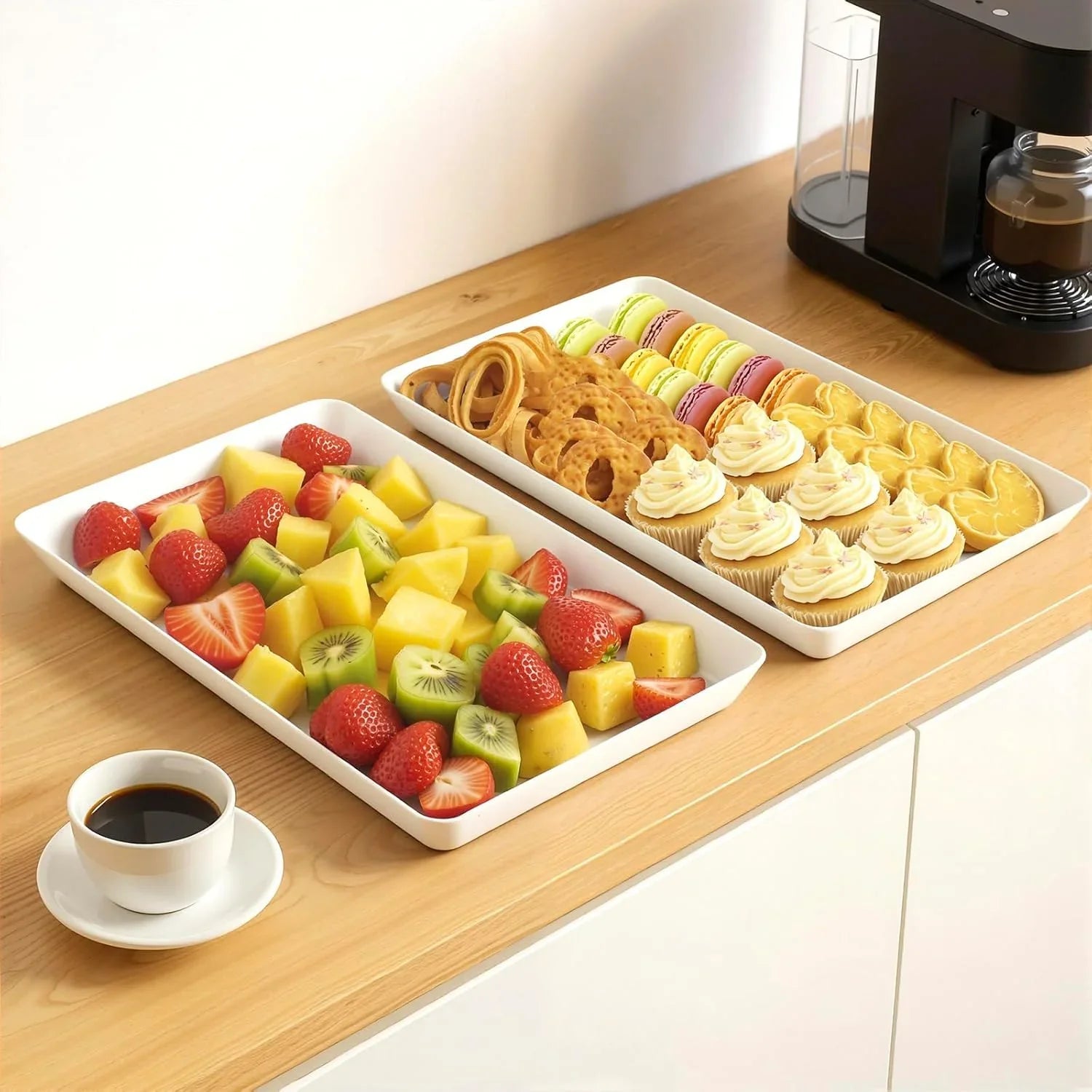 Two white serving trays on wooden counter with assorted fresh fruit and variety of desserts near a coffee cup and black coffee maker