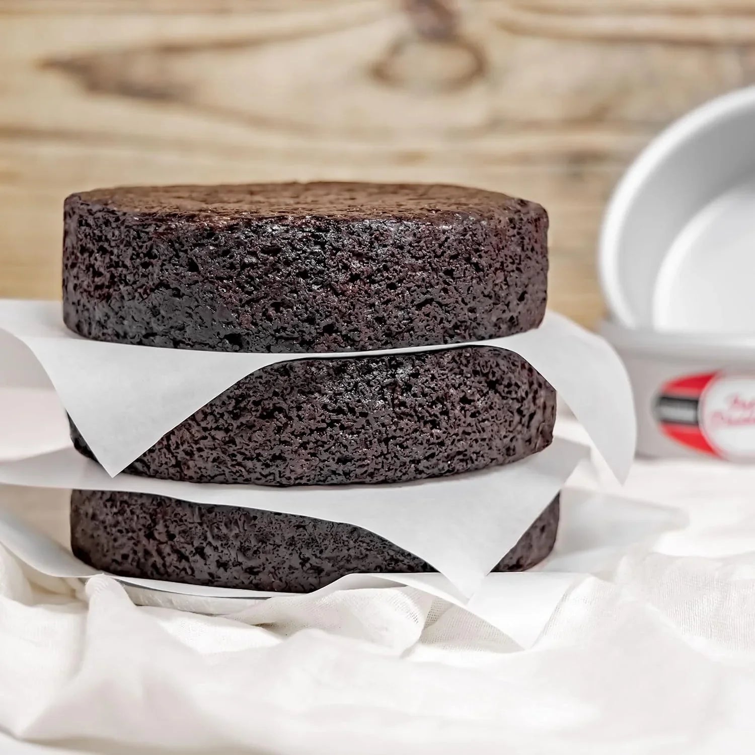 Three layers of moist chocolate cake separated by parchment paper stacked on white cloth