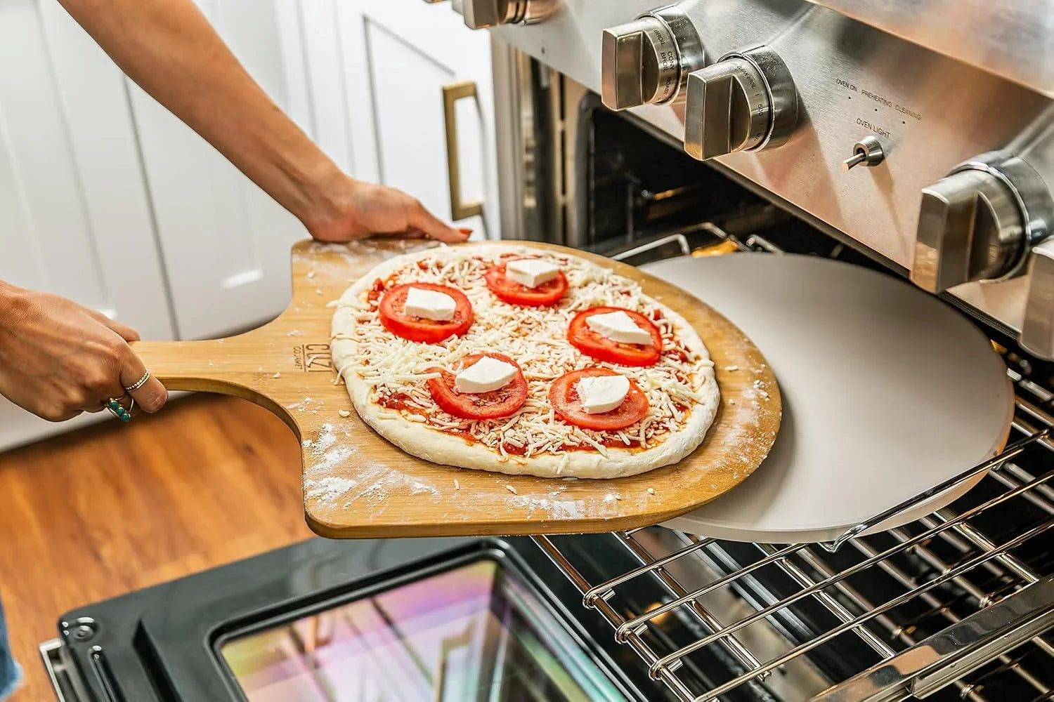 Person sliding raw pizza with tomato, cheese on bamboo peel into oven with stone baking surface