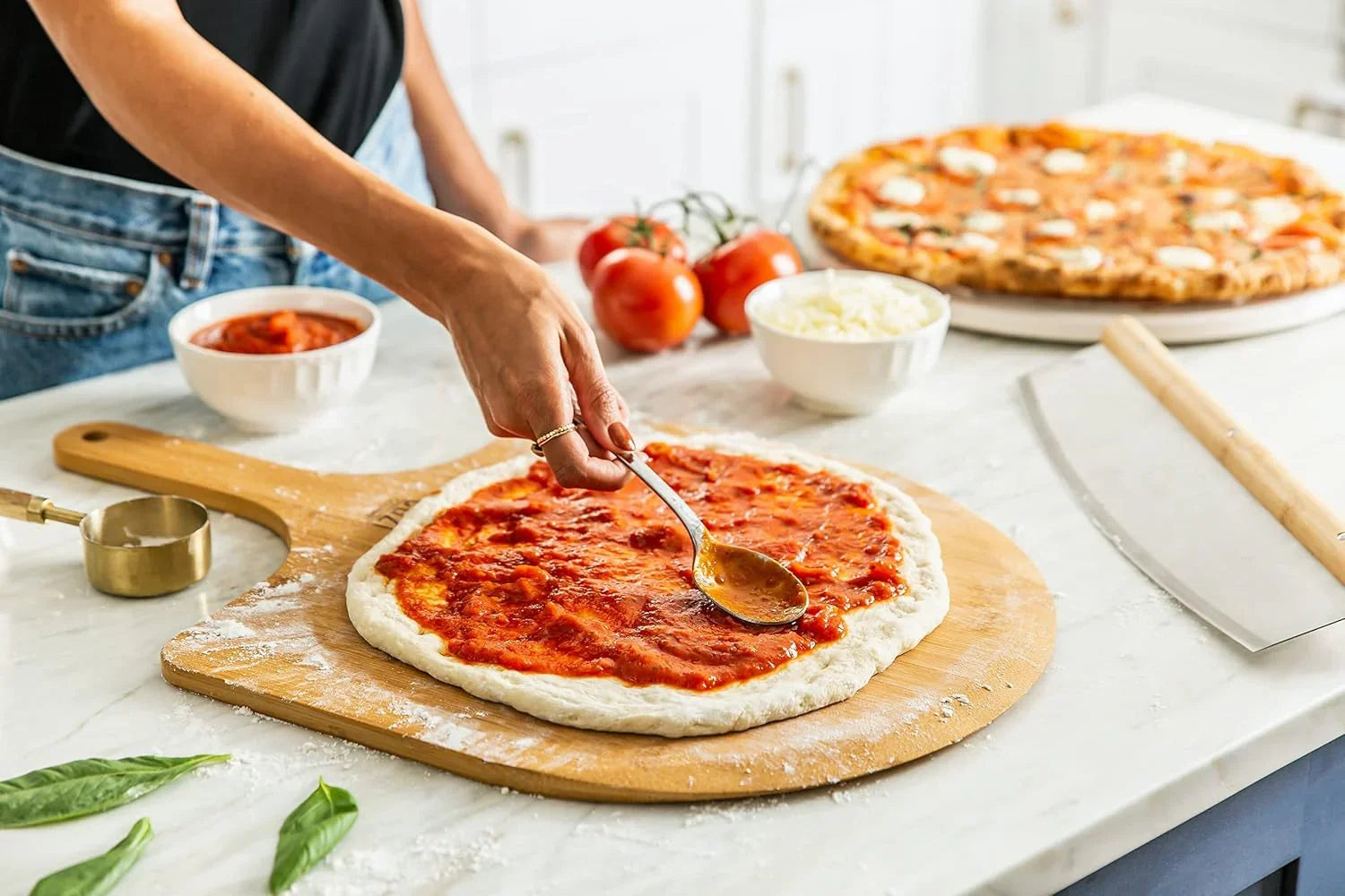 Hand spreading tomato sauce on pizza dough on wooden peel with toppings and cooked pizza in kitchen