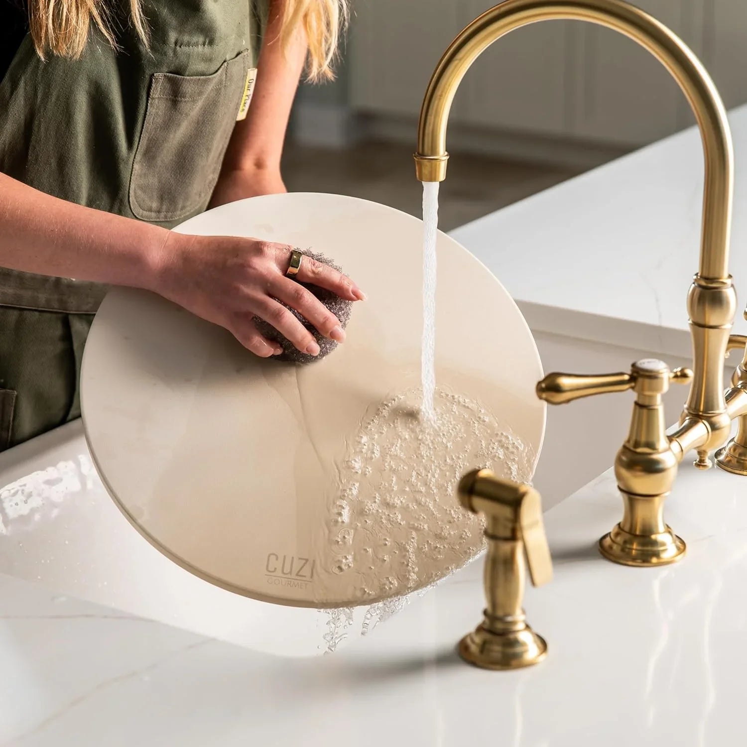 Person washing a large beige CUZIA Gourmet pizza stone under running water in a kitchen sink with brass faucet