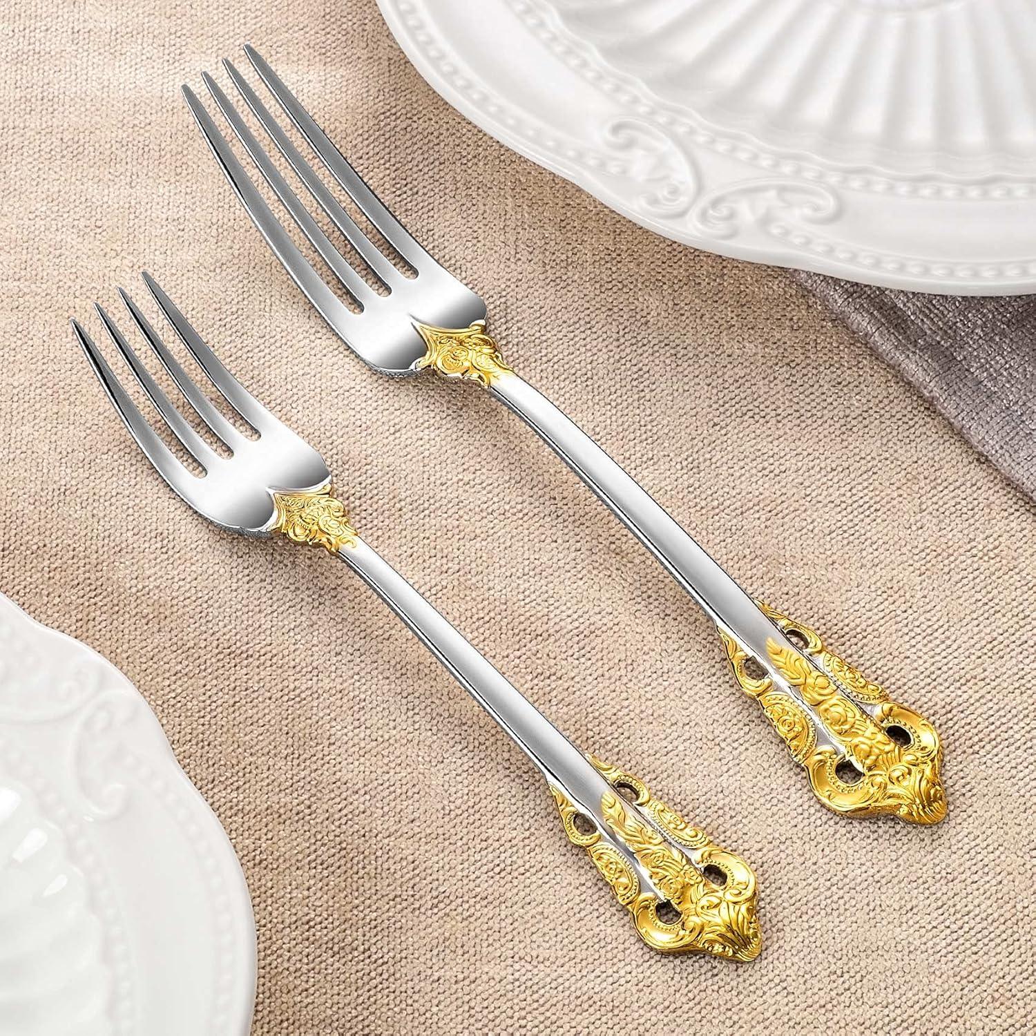 Two ornate silver forks with gold floral accents on handles, placed on beige fabric next to white embossed plates