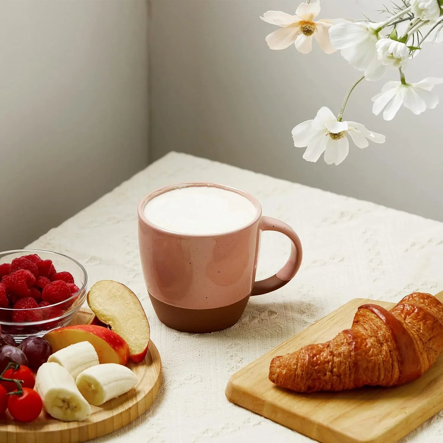 Pink and brown ceramic coffee mug with foam, croissant on wooden board, fresh fruit and flowers