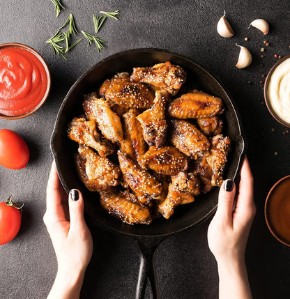 Hands holding cast iron skillet with sesame chicken wings surrounded by garlic, sauces, tomatoes, and rosemary