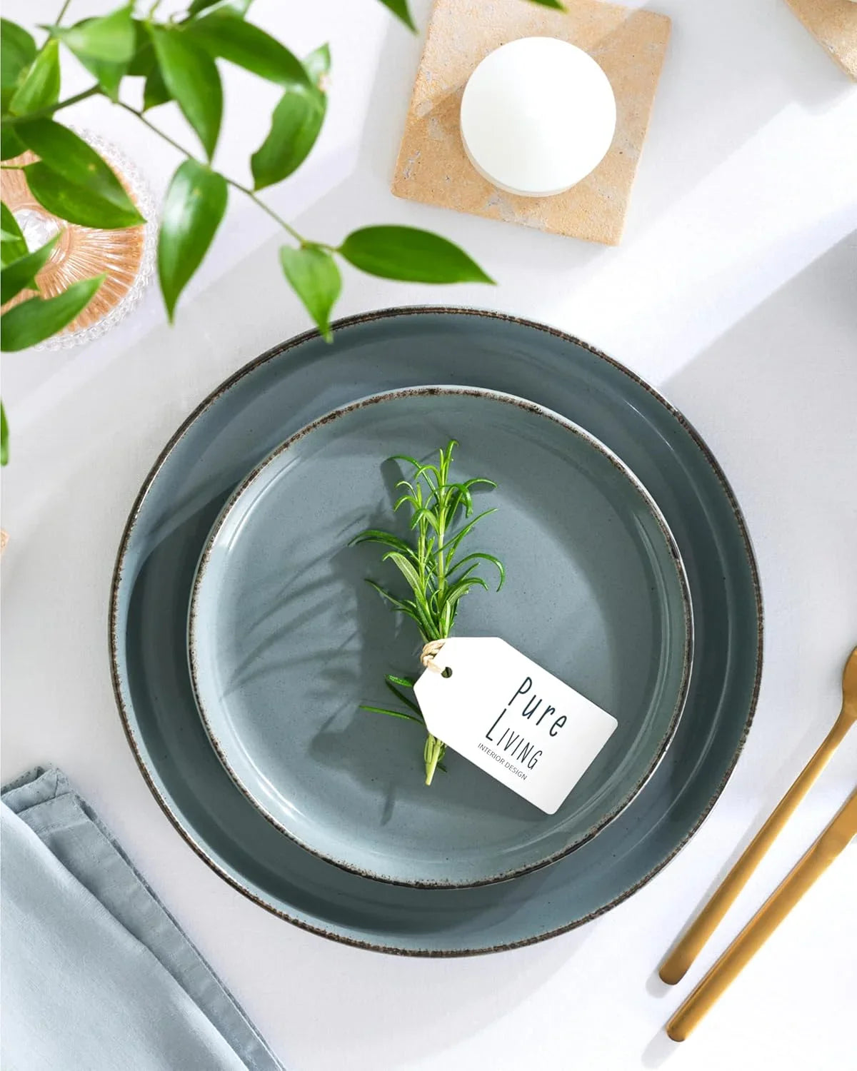 Rustic smoky blue stoneware dinnerware set with green sprig and Pure Living tag on white tablecloth