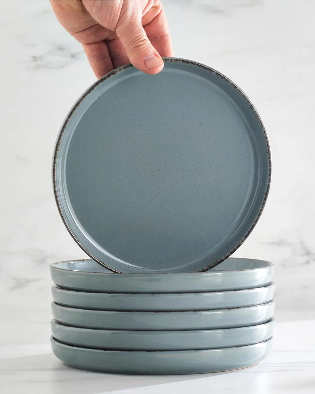 Hand holding rustic smoky blue stoneware plate above stack of matching plates on marble surface