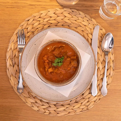 Top view of tomato-based stew in ceramic bowl on speckled plate with woven natural placemat and cutlery