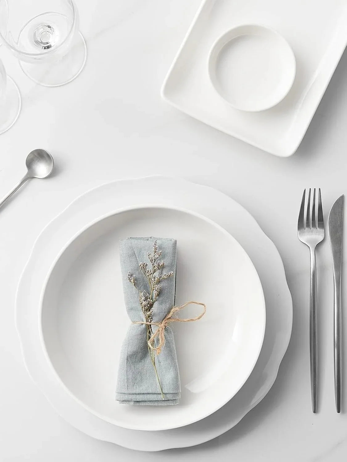 Minimalist white table setting with wide shallow porcelain pasta bowl, silver cutlery, blue linen napkin tied with twine and dried flowers